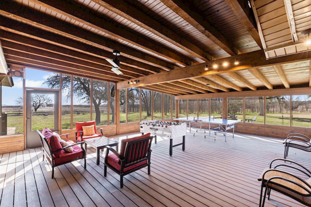 A spacious sunroom with a wooden ceiling and floor, featuring red cushioned seating, a foosball table, and ping pong table, offers outdoor views.