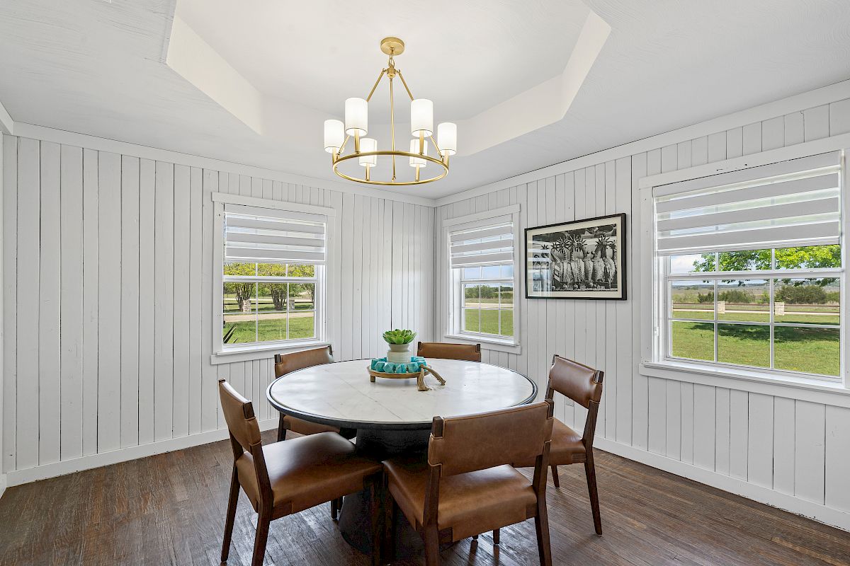 A dining room with a round table, four chairs, chandelier, and two windows. A framed picture hangs on the wall, offering a scenic view.