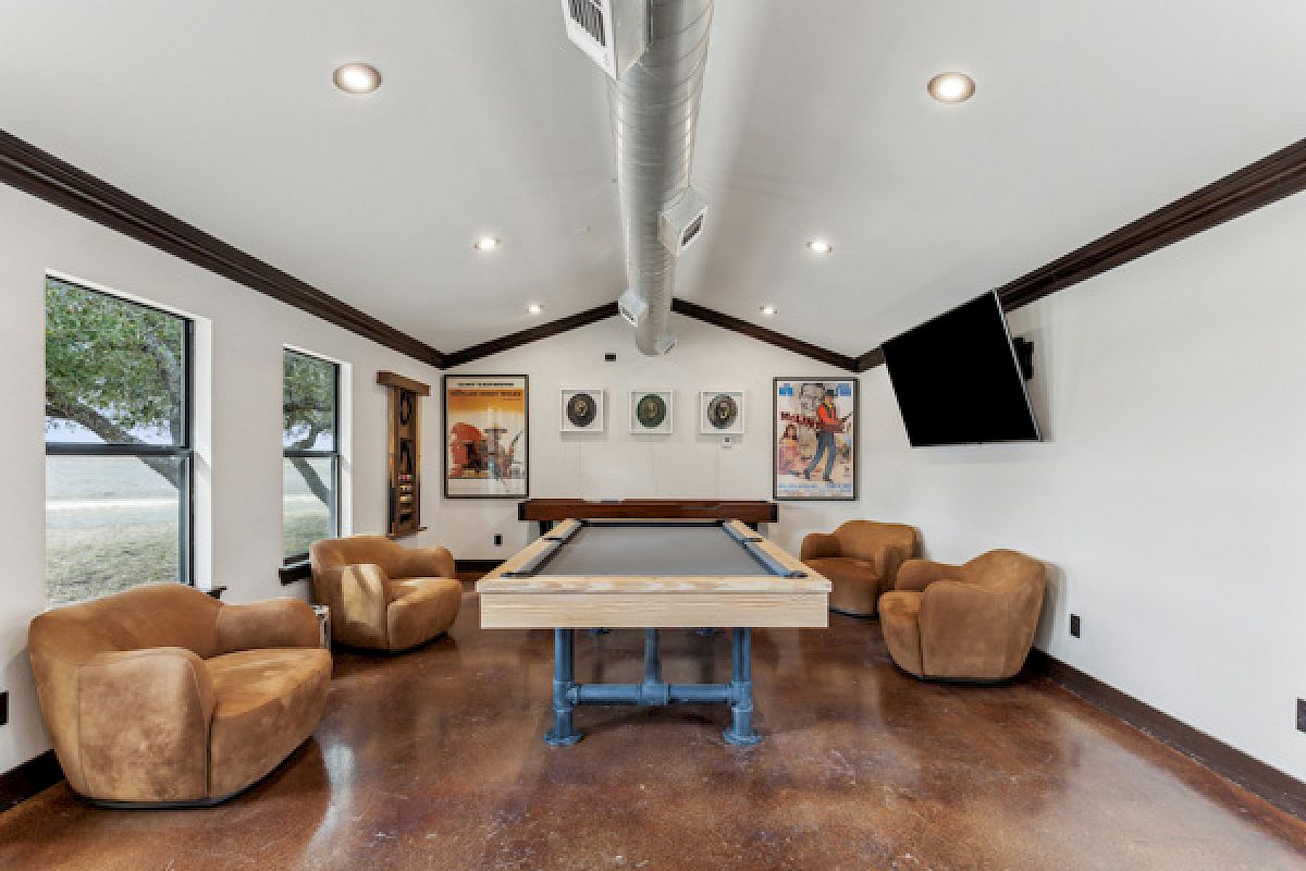 A game room with a pool table, four brown chairs, framed art, a TV, and large windows providing a bright and airy atmosphere.