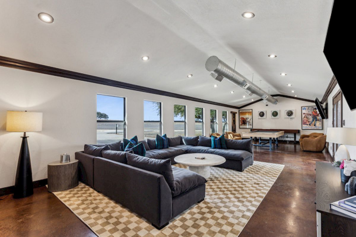 Spacious living room with large sectional sofa, round table, multiple windows, and wall artwork. Wood floors, ambient lighting.