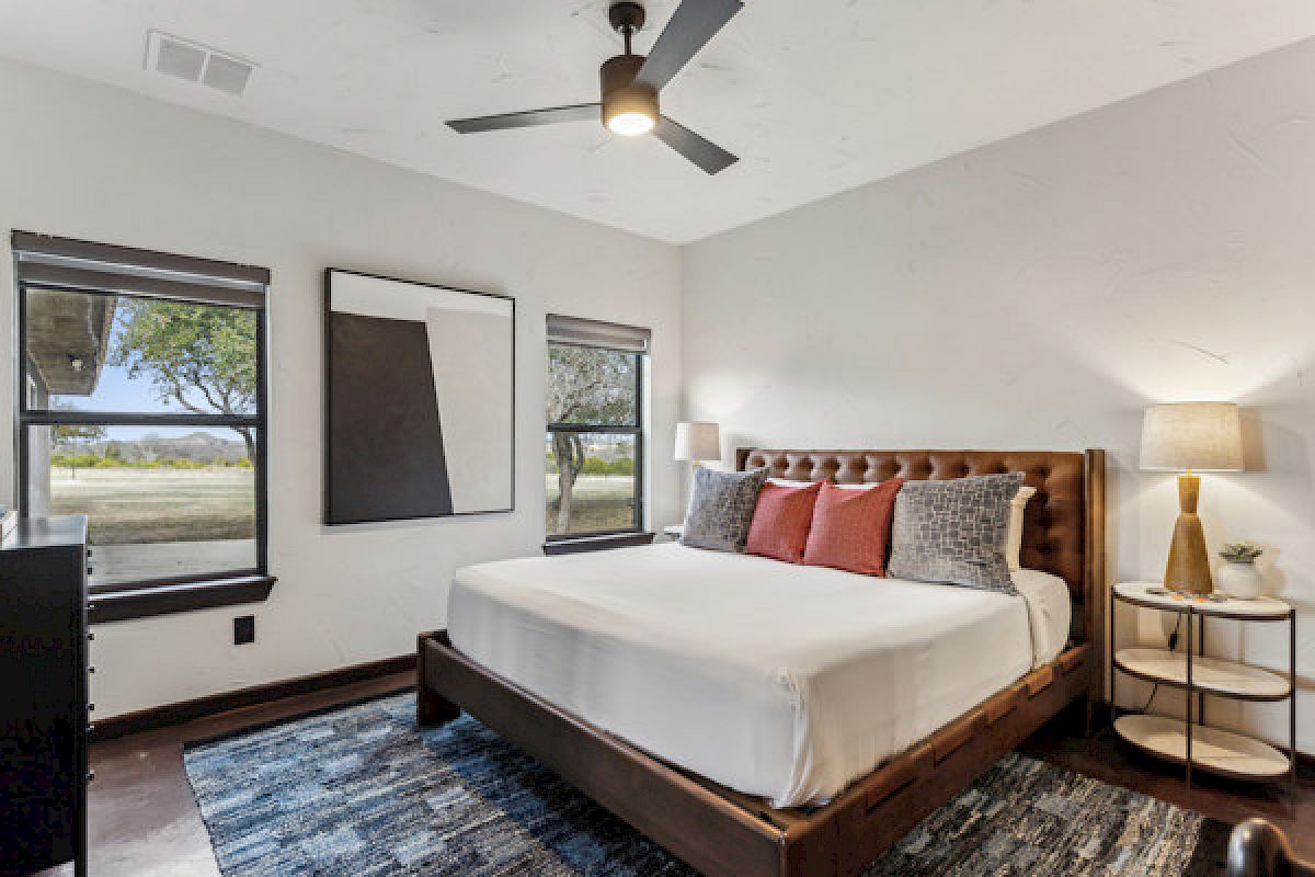A cozy bedroom with a large bed, decorative pillows, wall art, two windows, table lamp, ceiling fan, and a blue patterned rug.