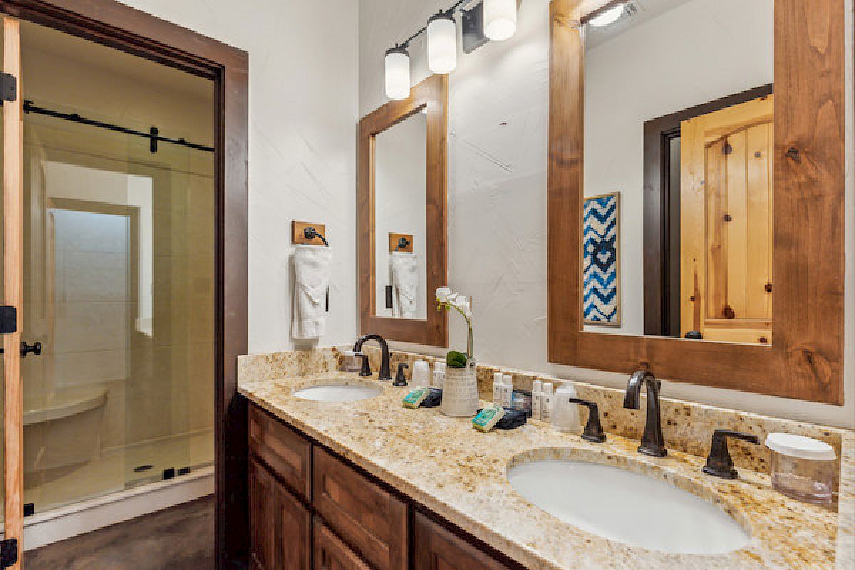 A bathroom with double sinks, granite countertops, wooden cabinets, mirrors, and a shower with a glass door.
