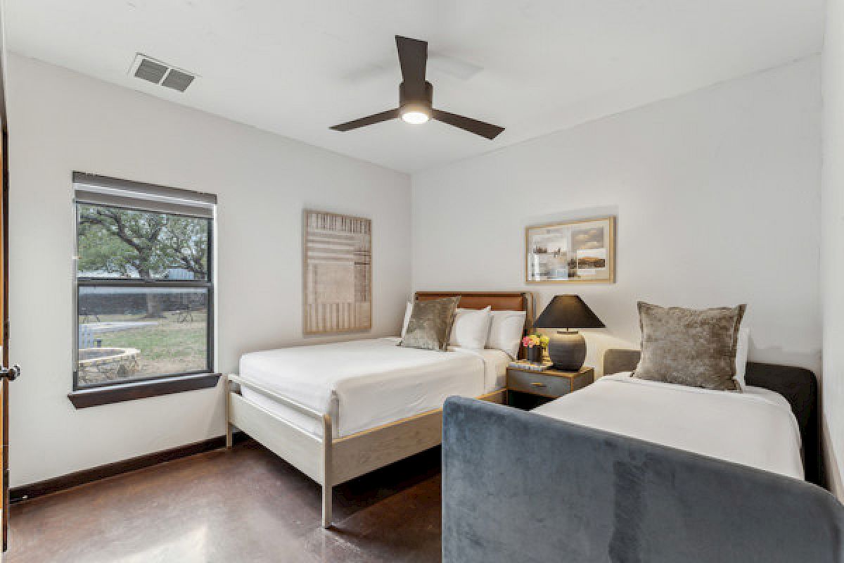 A bedroom with a double bed and a single bed, wall art, a ceiling fan, a lamp, and a window showing outdoor greenery.