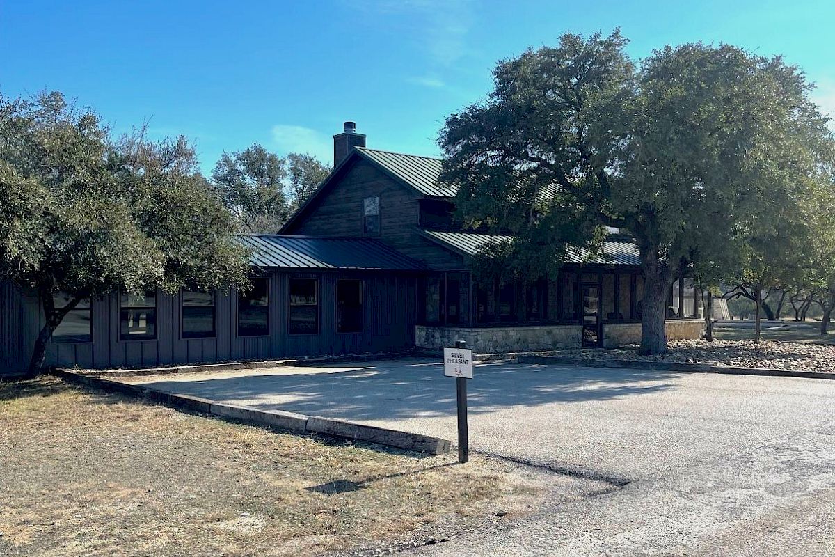 A rustic cabin with a tin roof surrounded by trees, set in a spacious area with a paved path leading to it. The sky is clear and blue.