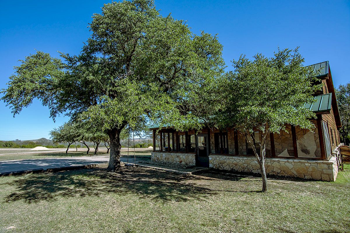 A rustic stone building with a green roof sits under large, leafy trees on a sunny day, with a pathway and lawn surrounding it.