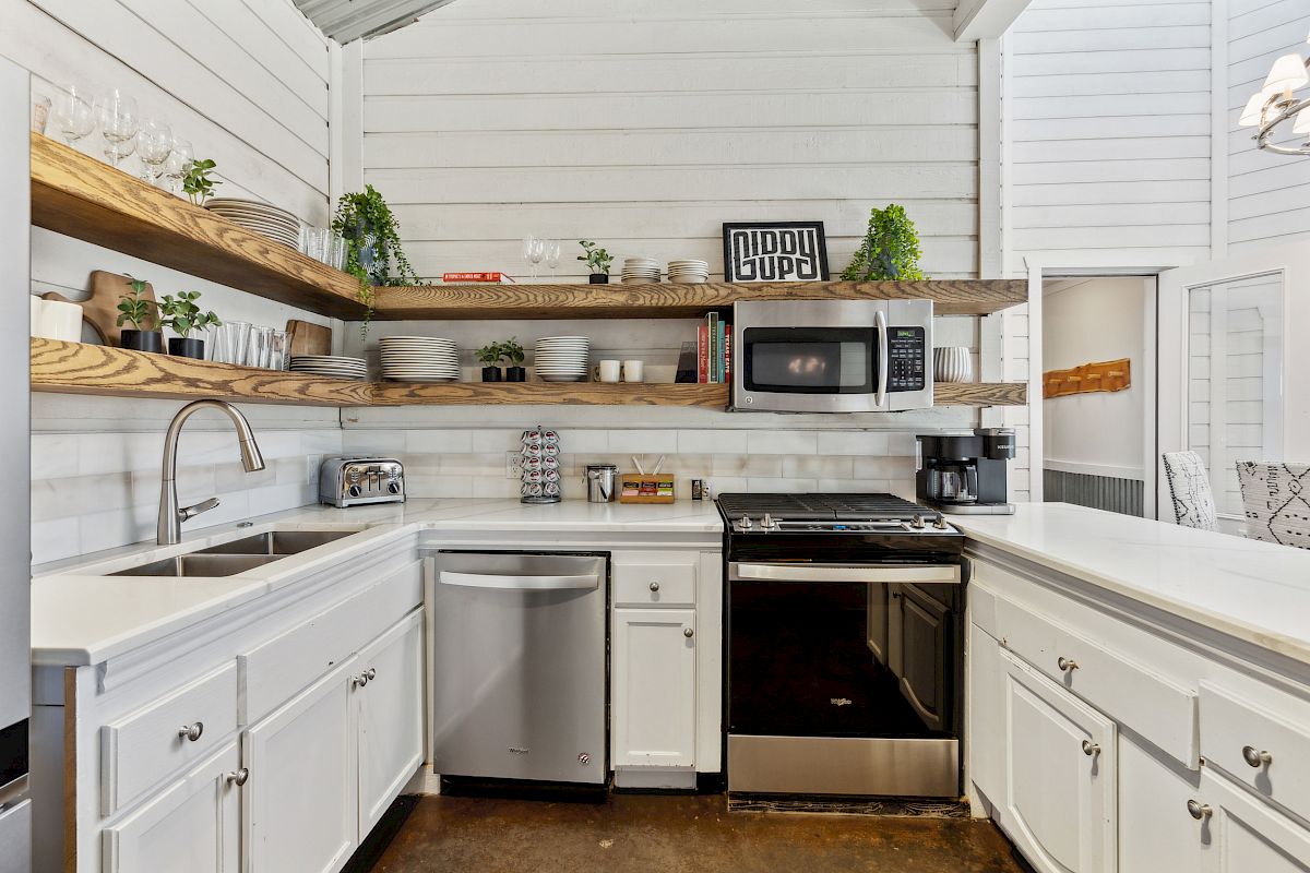 A modern kitchen with white cabinets, open wooden shelves, stainless steel appliances, and decorative plants on display.