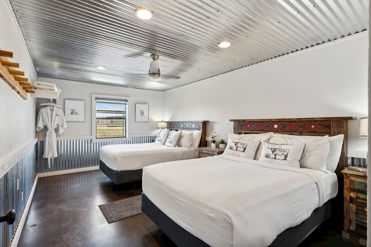 A rustic bedroom with two beds, metal ceiling, white robes, and framed pictures on white walls, featuring decorative wooden headboards.
