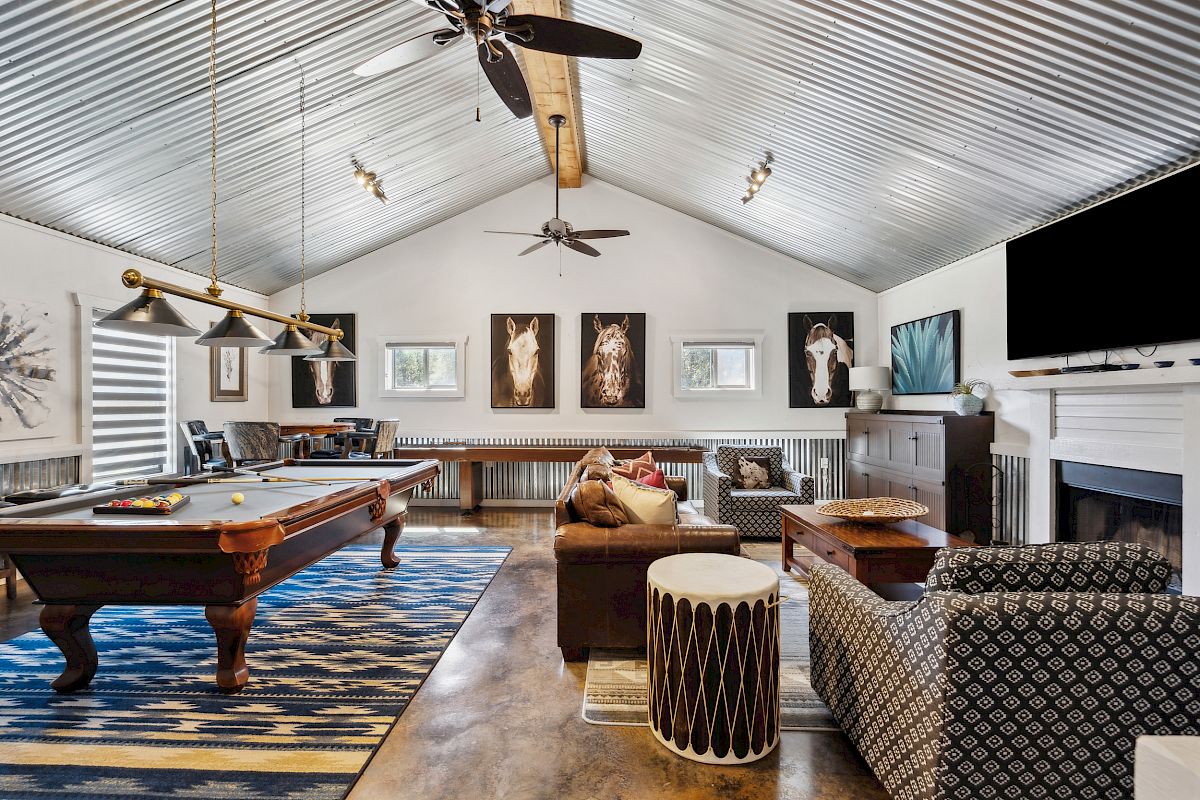 A stylish game room with a pool table, seating area, and a TV on the wall. Decor includes paintings, and the ceiling is metallic.