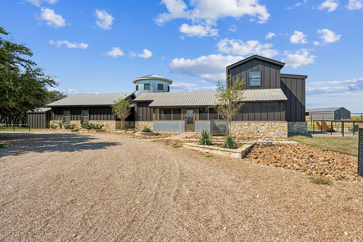 The image shows a rustic building with a stone base and metal siding, surrounded by a gravel driveway and sparse landscaping.