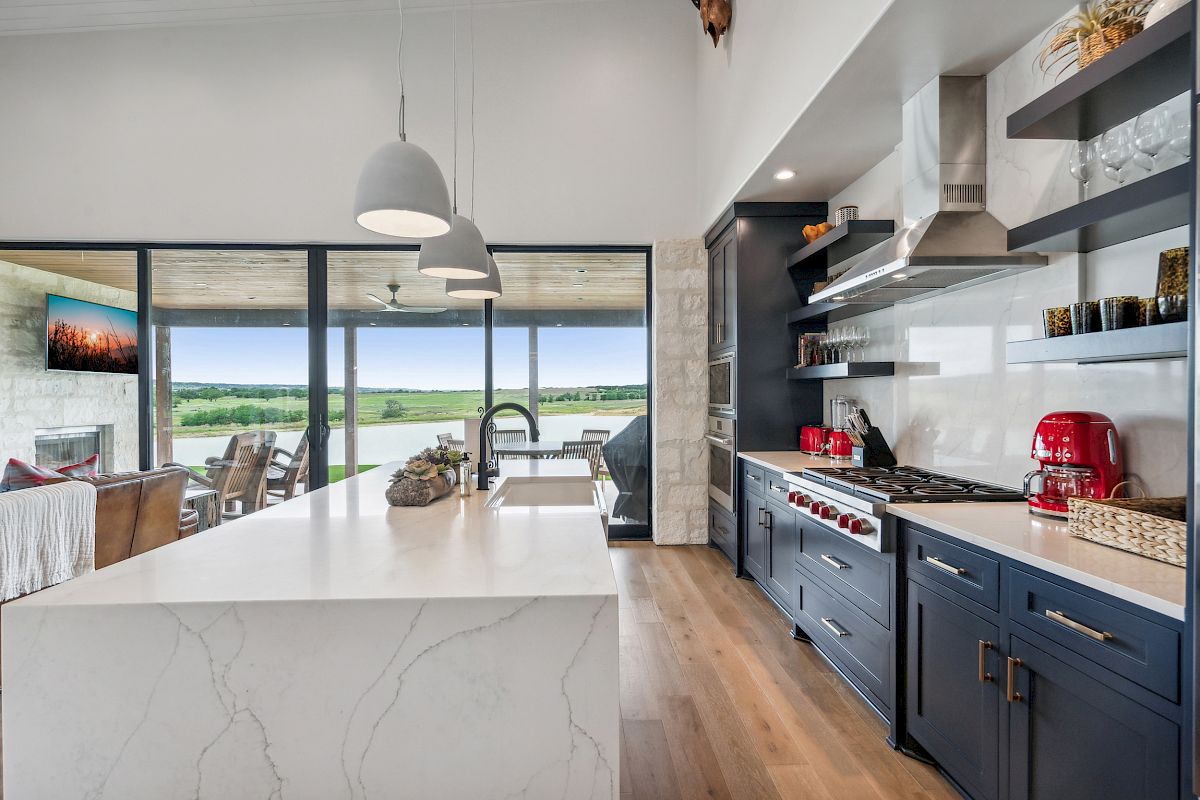 A modern kitchen with a marble island, sleek cabinets, and large windows overlooking a scenic view, featuring a red kettle and stove.