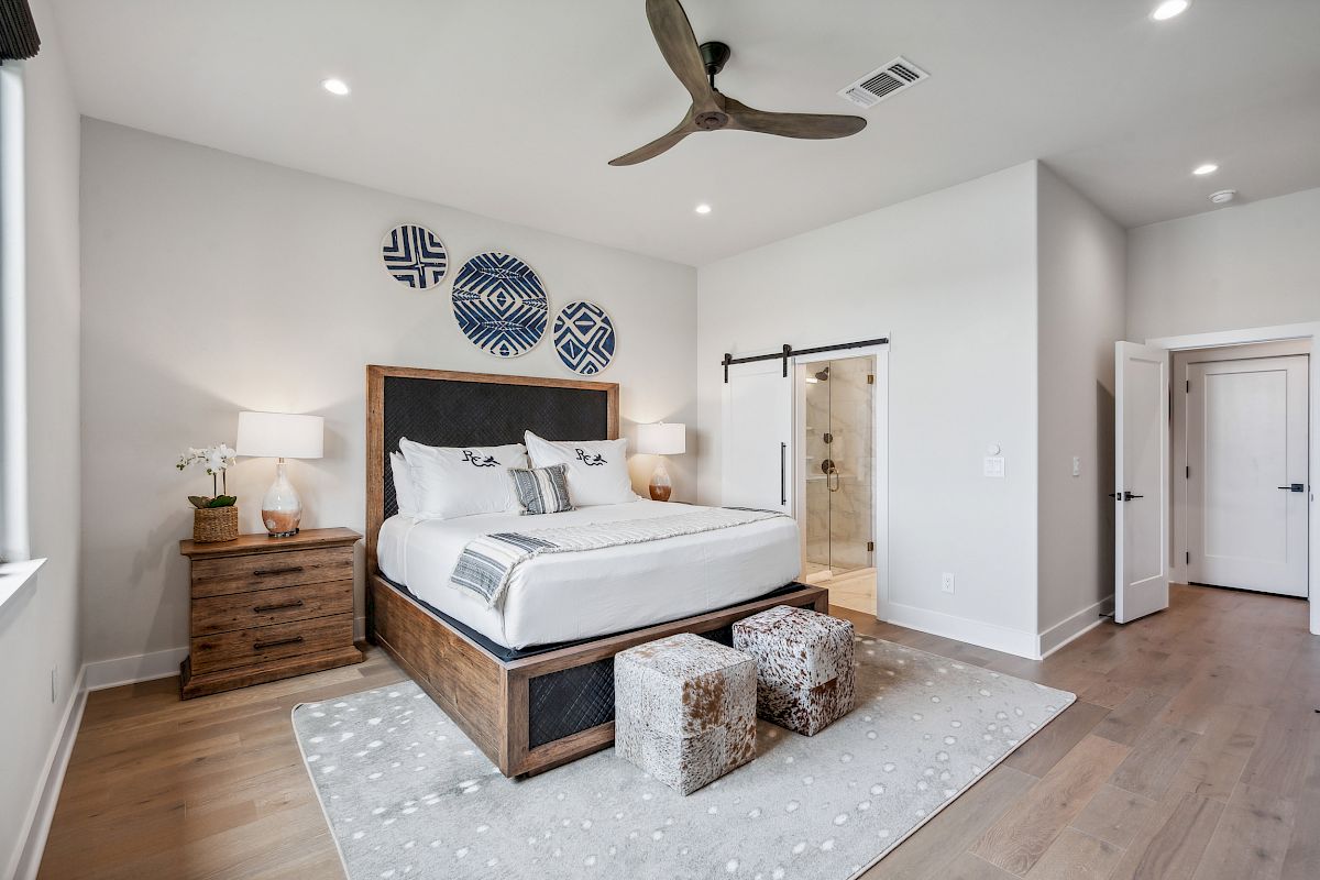 A bedroom features a large bed, bedside tables with lamps, wall art, and a doorway to a bathroom, all with a modern decor style.