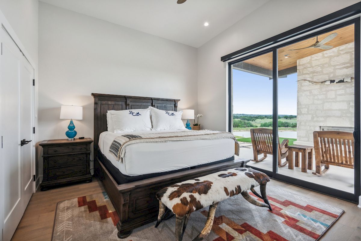 A bedroom with a large bed, cowhide bench, lamps, and glass doors opening to a patio with chairs overlooking a scenic outdoor view.