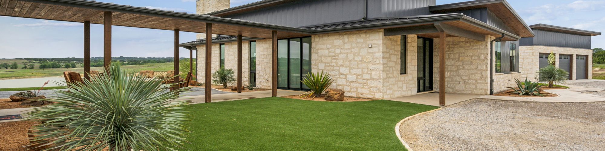 A modern house with stone facade, large windows, and a pathway surrounded by a well-maintained lawn under a blue sky with clouds.