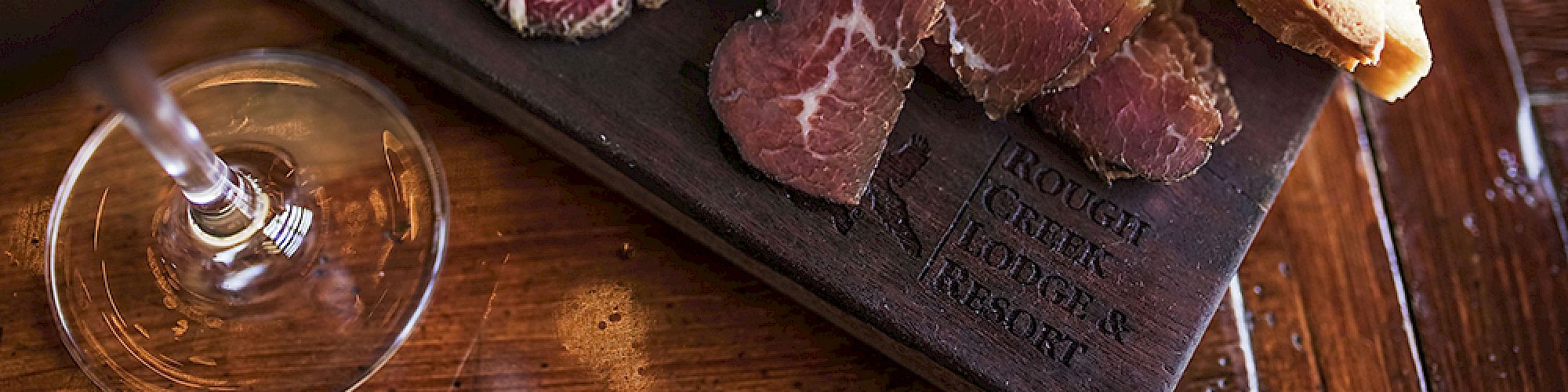 This image shows a charcuterie board with sliced meats, pickles, and toast, along with a glass of red wine on a wooden table.