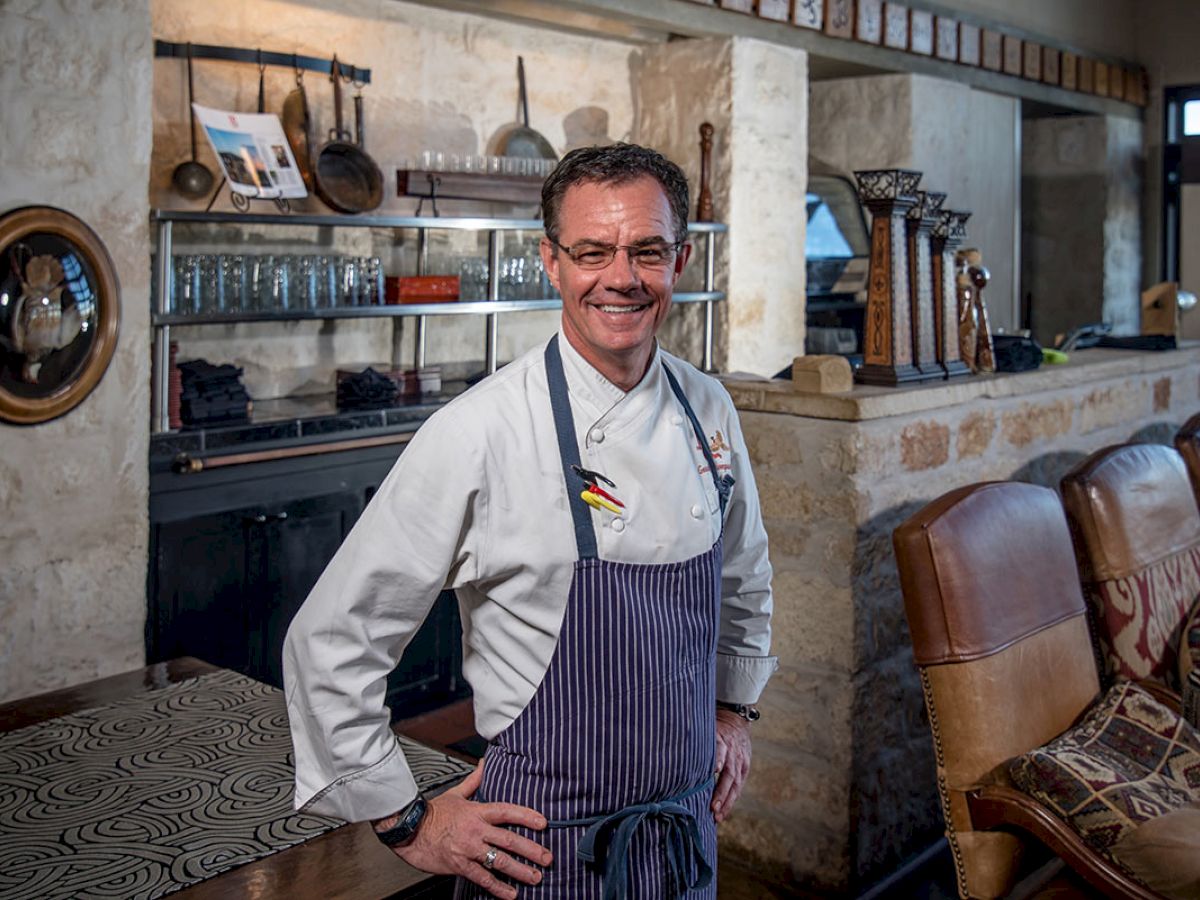 The image shows a chef dressed in a uniform and apron, standing in a rustic kitchen or restaurant setting, smiling at the camera.