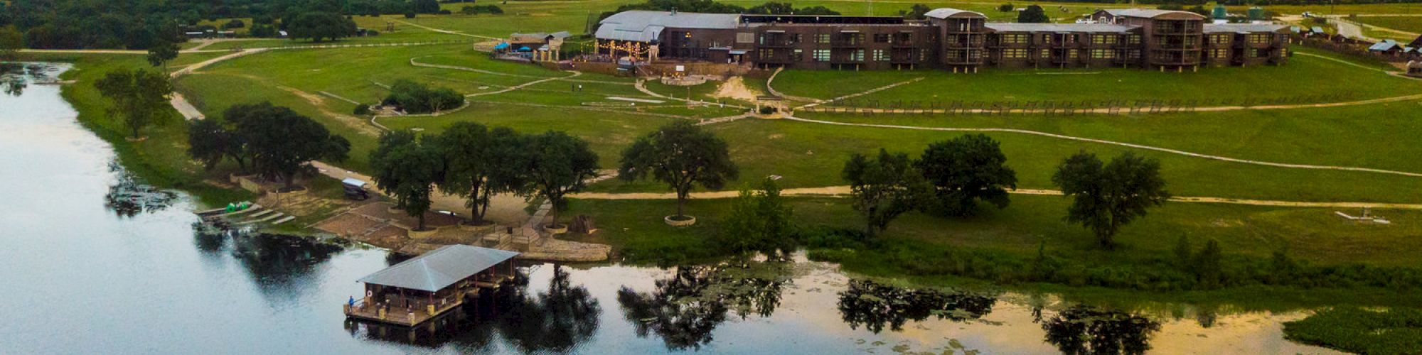 A large building complex is situated by a serene lake, surrounded by green lawns, trees, and pathways, featuring a docked structure on the water.