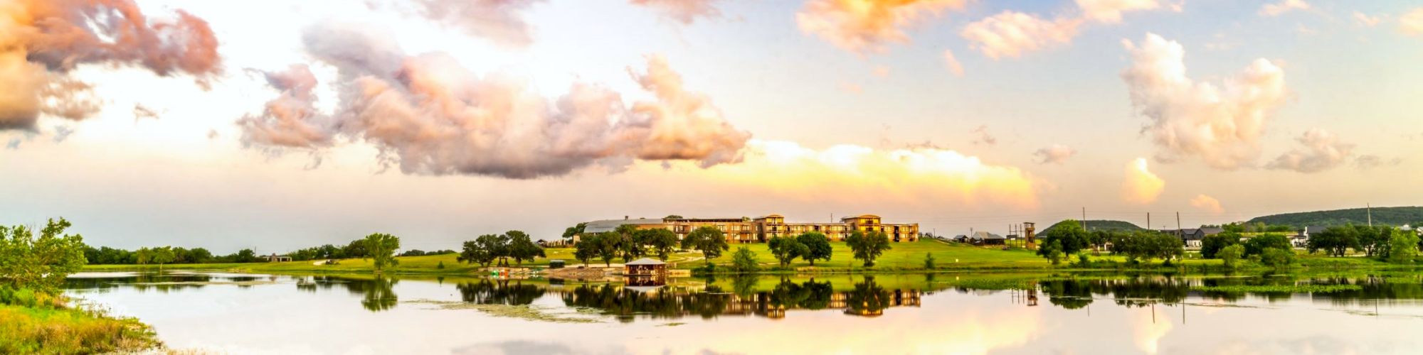 A serene landscape showing a lake with a grassy shoreline, reflecting a vibrant sky with clouds and distant buildings under the colorful sky.