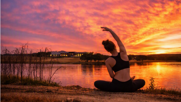 A person does yoga in a black outfit by a lakeside at sunset with dramatic orange and pink clouds, taking a peaceful pose.