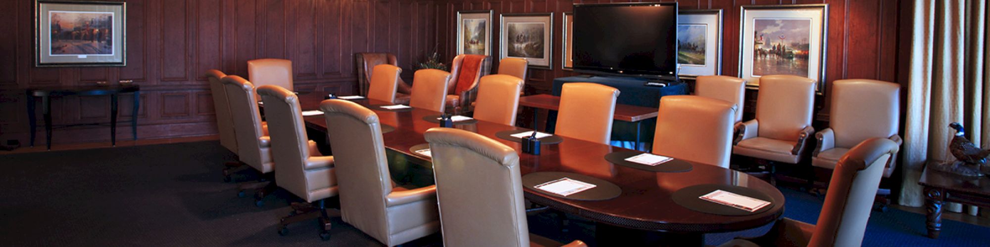 A conference room with a long table, twelve chairs, a TV, and framed pictures on wooden-paneled walls, illuminated by ceiling lights and chandeliers.