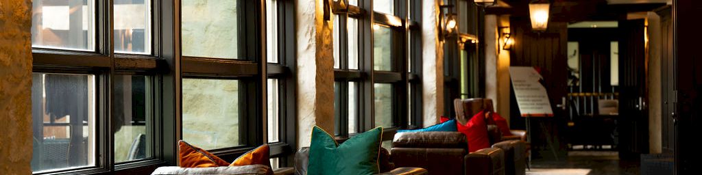 A cozy hotel hallway with leather sofas, colorful cushions, warm lantern lights, and large windows along the corridor.