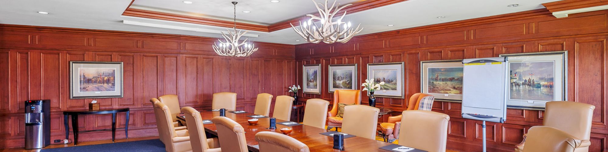 A conference room with a long table, leather chairs, artwork on the wood-paneled walls, and a flip chart stand.