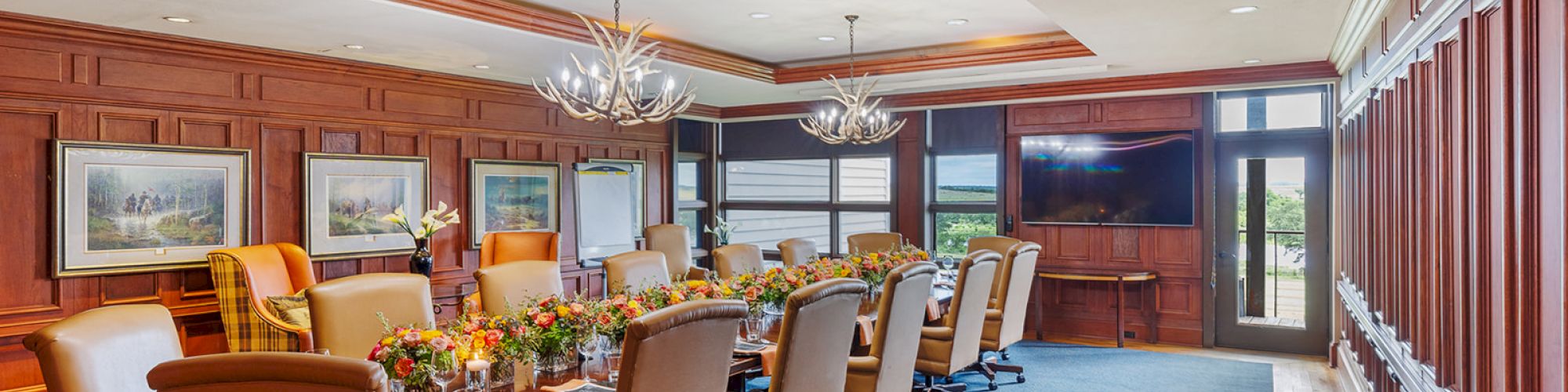 A formal conference room with a long table, leather chairs, chandeliers, artwork on the walls, and a large TV screen at the back.