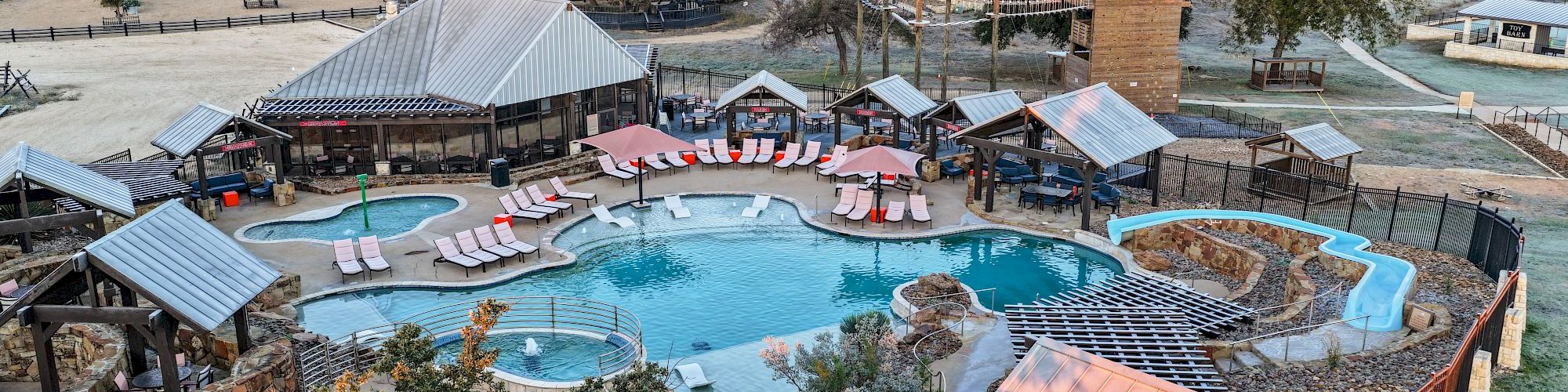 Aerial view of a ranch-style resort with a large turquoise pool, cabanas, lounge chairs, and surrounding cabins.