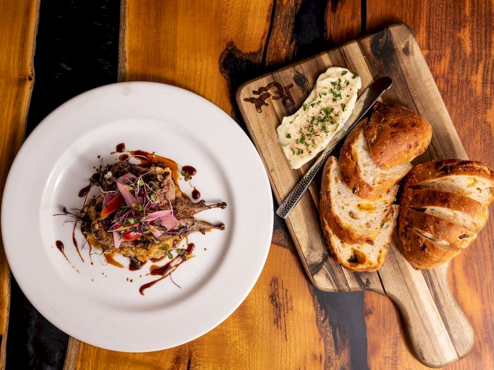 A plated entr&eacute;e with garnishes and drizzle sits beside sliced toasted bread and herb butter on a wooden board.