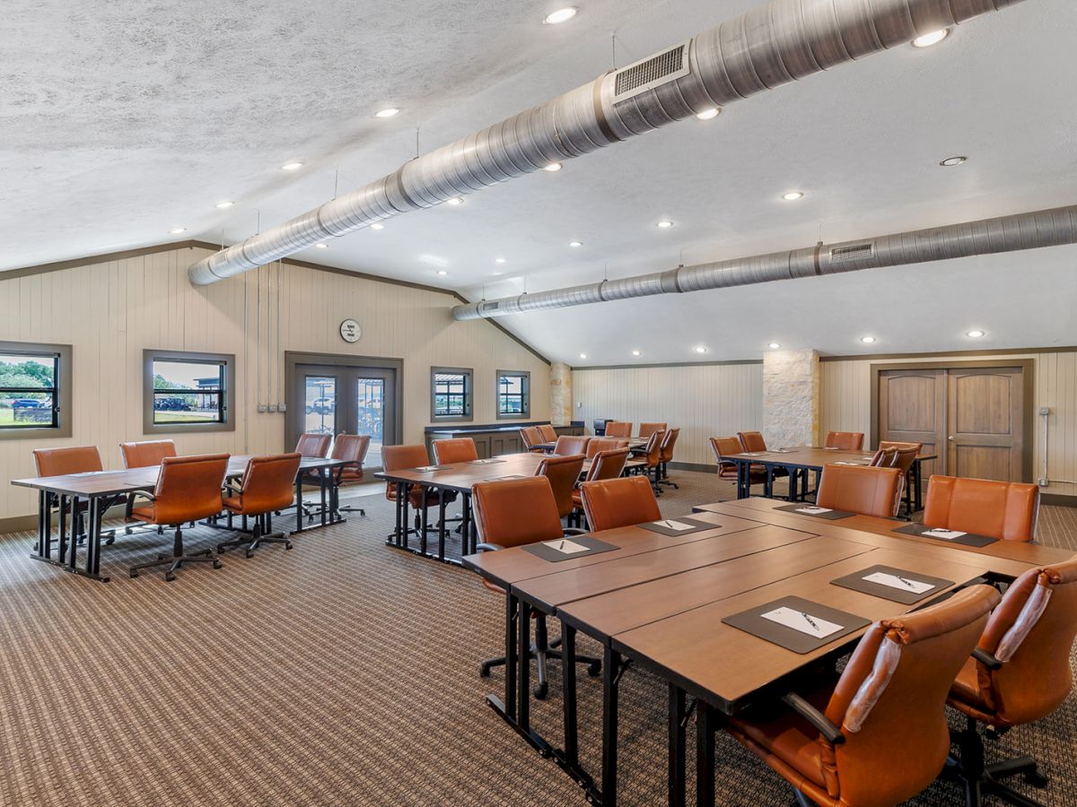 A conference room with tables, brown chairs, and notepads, under a ceiling with exposed ductwork and framed pictures on the walls.