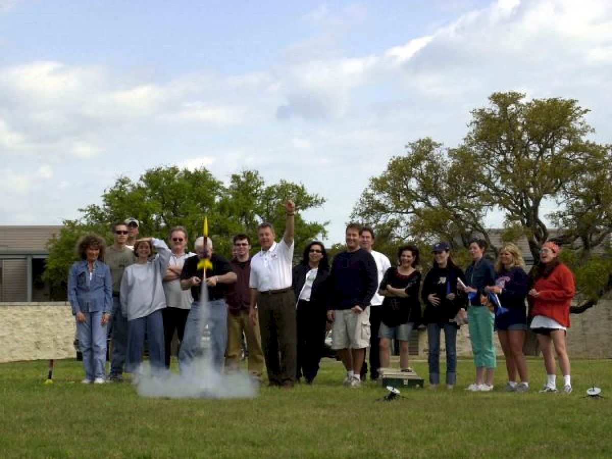 A group of people outdoors, some cheering as a model rocket launches with smoke around it, and trees and a building visible in the background.