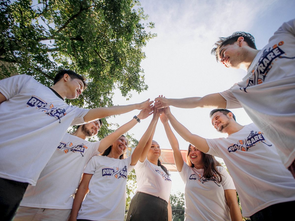 A group of people in matching white shirts, standing in a circle, are raising their hands together in a team gesture of unity outdoors.