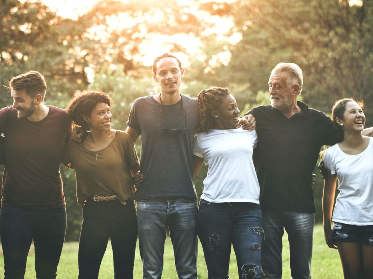 A diverse group of people are standing outside, arms around each other, smiling, and enjoying the sunset in the background.