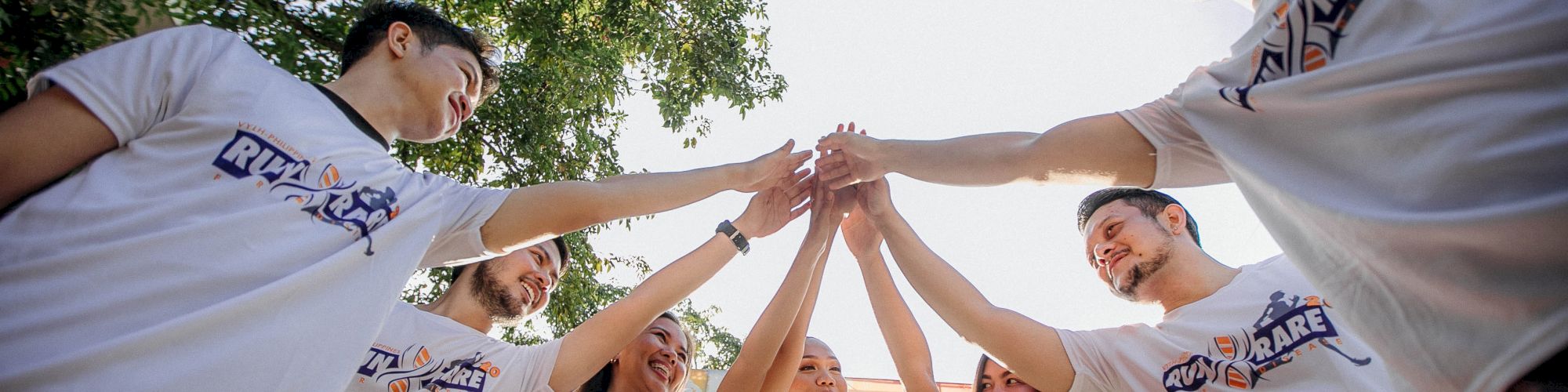 A group of people in matching white shirts is standing in a circle outdoors, touching hands in the center, smiling, and looking up.