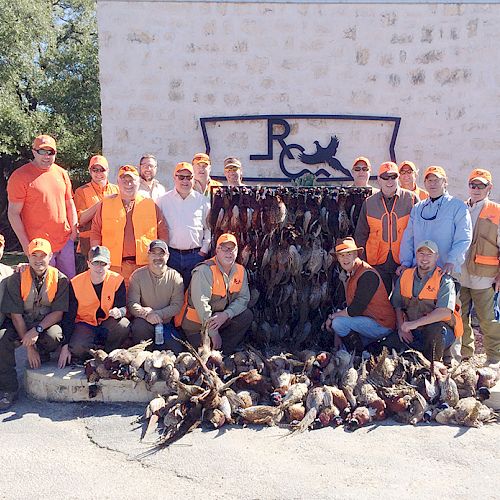 A group of people in hunting gear pose with their game birds in front of a wall featuring a logo of a leaping dog. The sentence ends.