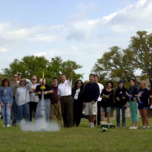 A group of people stands outdoors, watching and cheering as a model rocket launches, with smoke visible at its base.