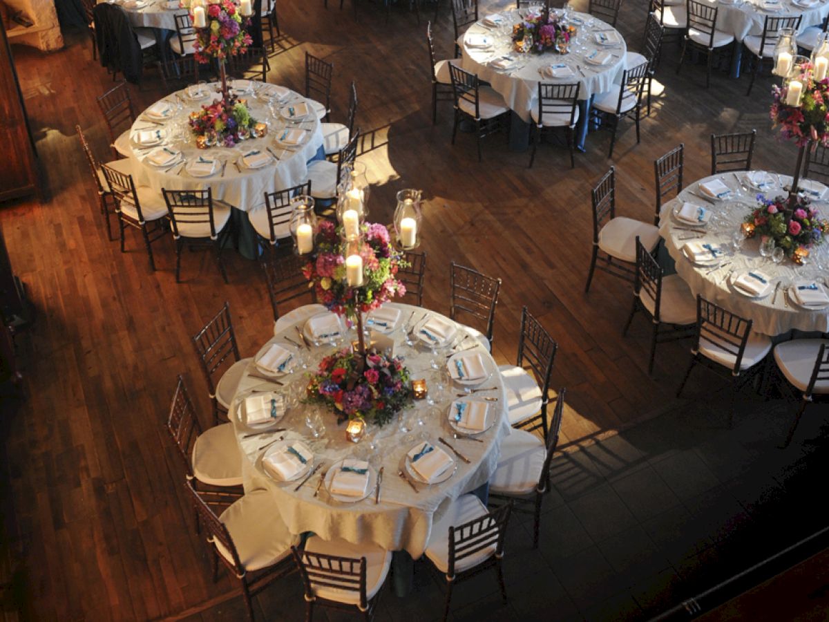 The image shows a beautifully set banquet hall with round tables, white tablecloths, floral centerpieces, and elegant place settings on a wooden floor.