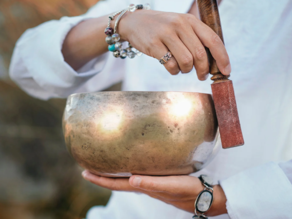 A person strikes a brass singing bowl with a mallet, wearing bracelets and a ring.
