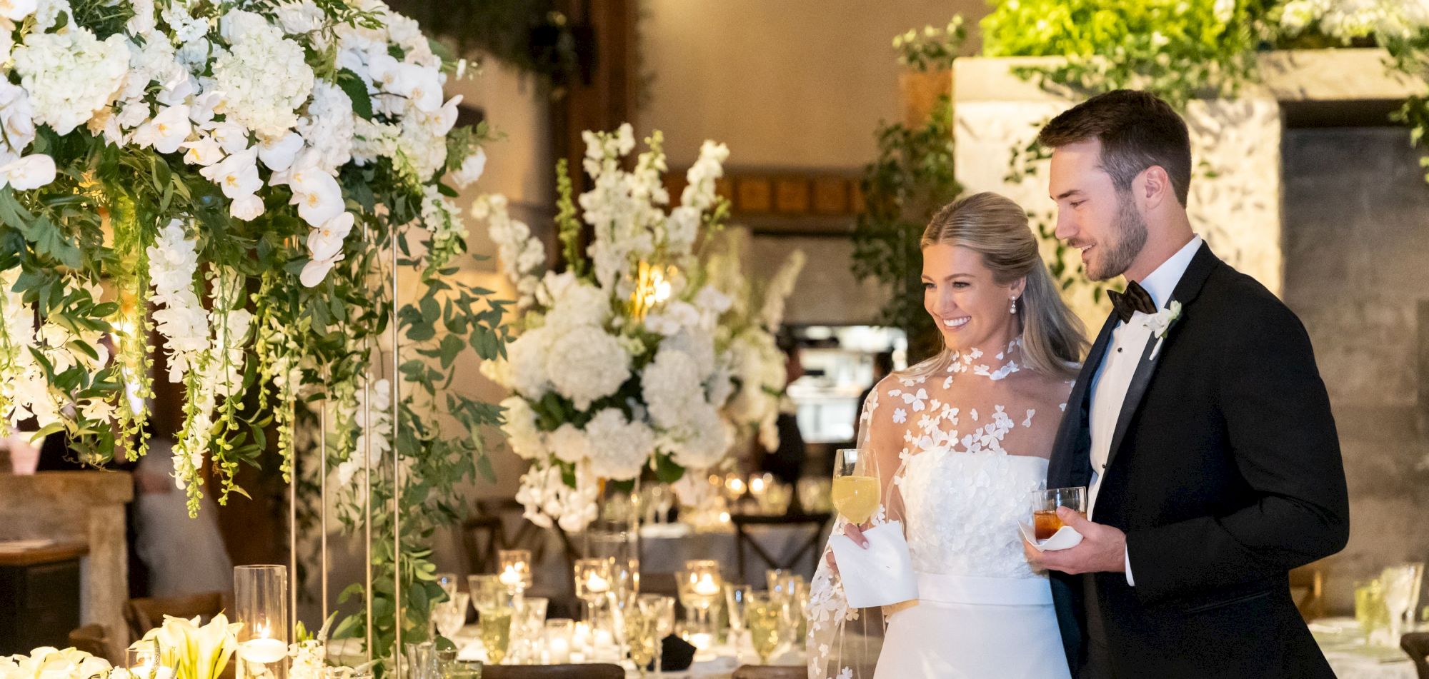 A couple at a wedding reception, surrounded by elegant floral arrangements and lit candles, with drinks in hand.