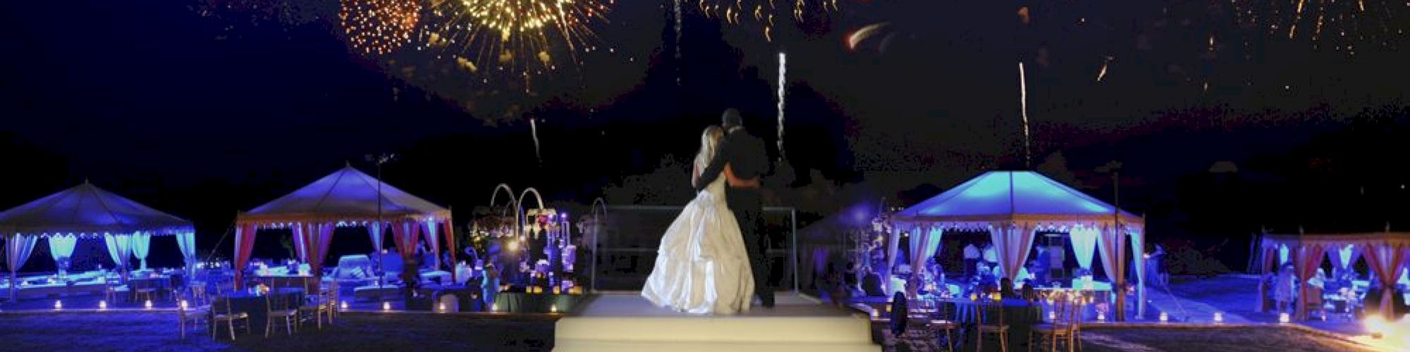 A couple in wedding attire stands on a platform, watching a vibrant fireworks display in the night sky, with decorated tents in the background.