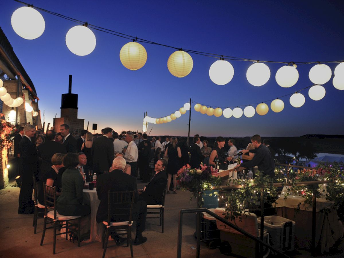 This image shows an outdoor evening gathering with hanging lanterns, people sitting at tables, and others standing and socializing under a dusk sky.