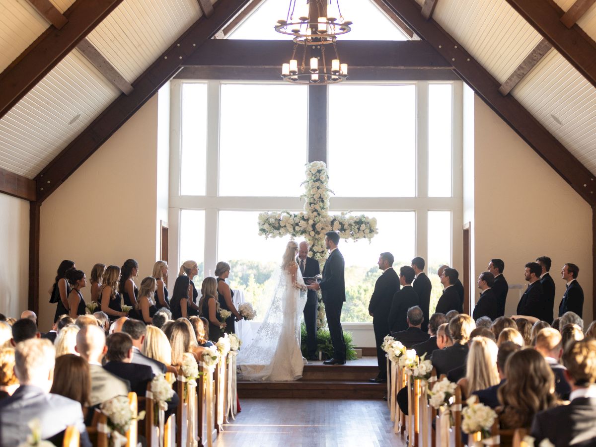 A wedding ceremony inside a chapel with a couple at the altar, surrounded by bridesmaids and groomsmen, facing the seated guests.