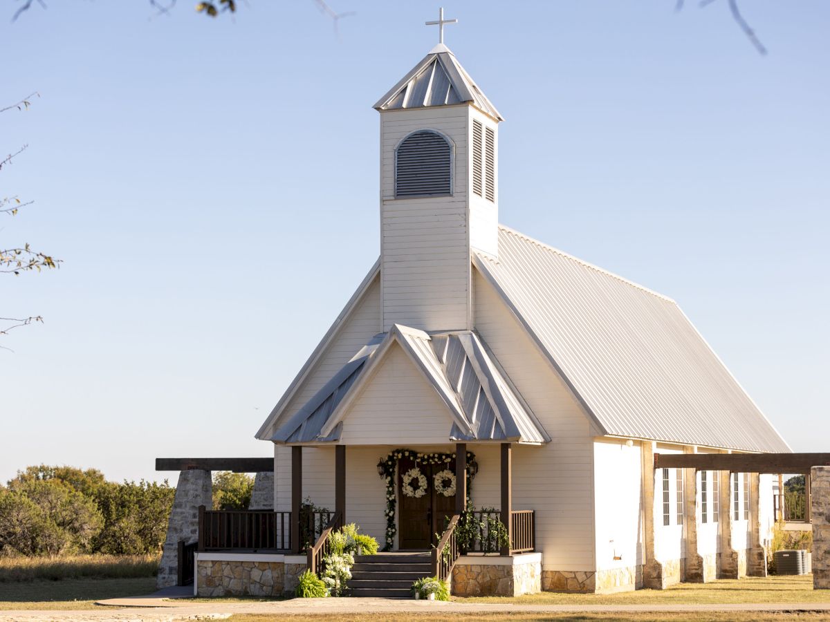 A small, white chapel with a cross on the steeple and rustic decor, situated in a countryside setting.
