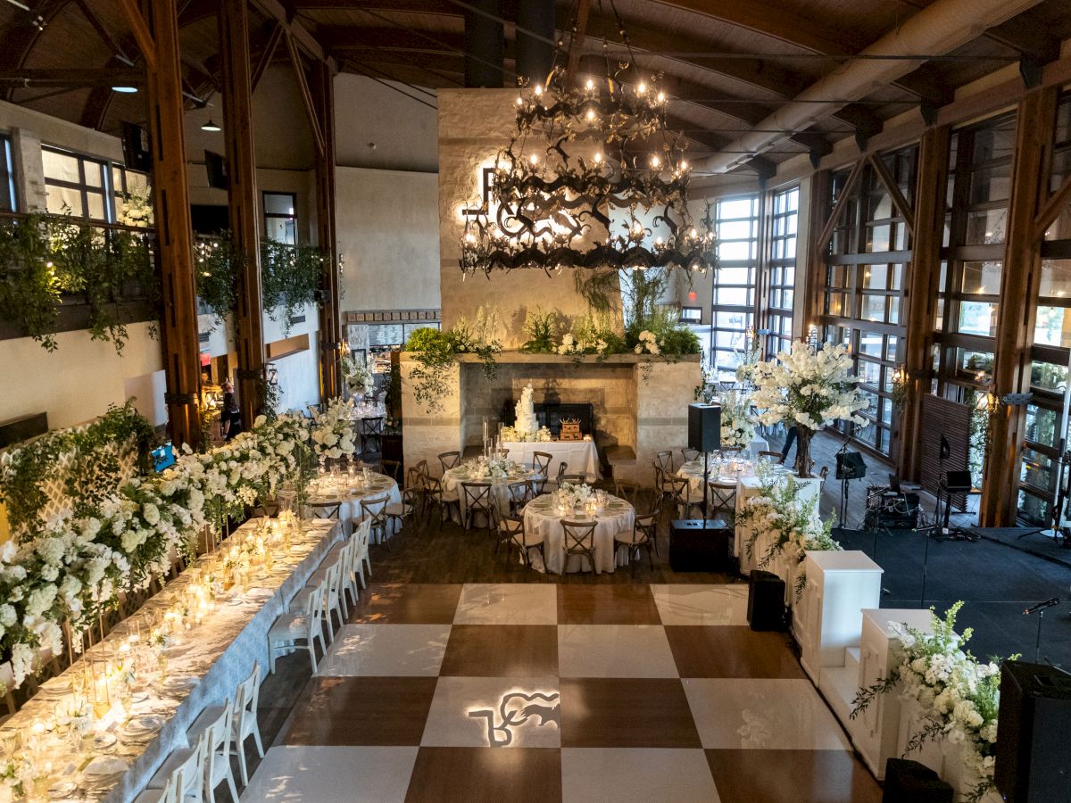 An elegant event space with floral decor, set tables, chandeliers, and checkered flooring, ready for a celebration or reception.