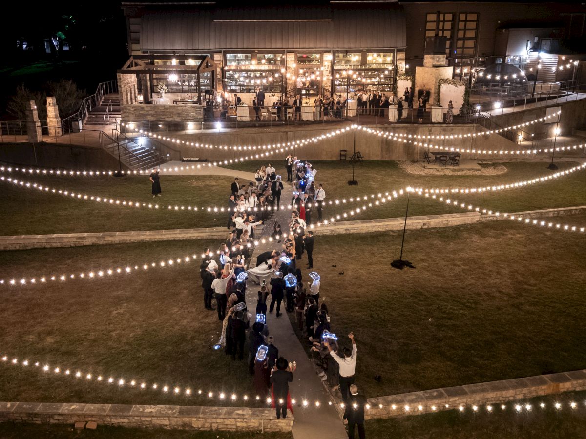 A festive outdoor night scene with string lights, a pathway of people celebrating, and a warmly-lit building in the background.