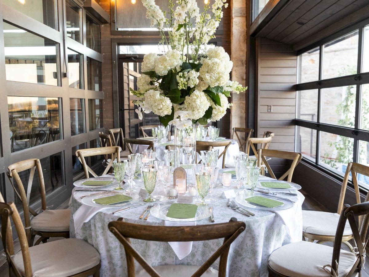 A beautifully set round table with elegant glassware, green menus, white floral centerpiece, and rustic wooden chairs, under warm lighting.