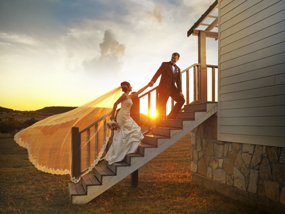 A bride and groom are walking down outdoor stairs at sunset, with the bride's veil flowing and the sun setting in the background.