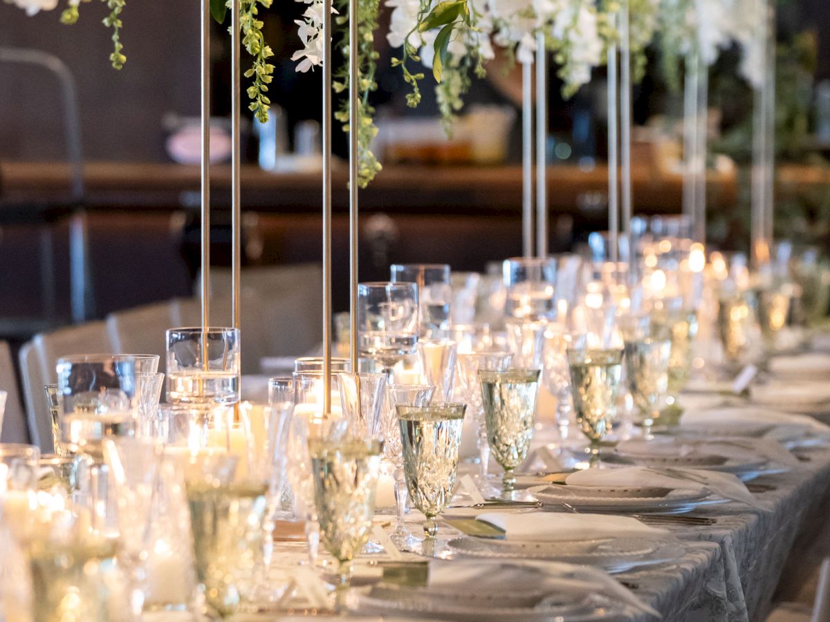 A beautifully set dining table with crystal glasses, plates, and elegant hanging white flower arrangements, softly lit by candles.