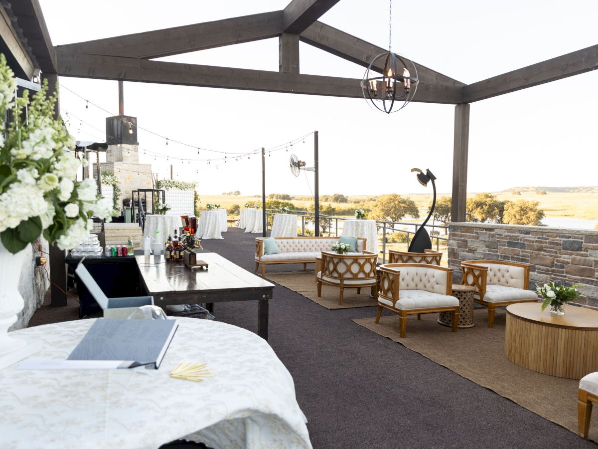 Outdoor event setup with lounge furniture, tables, flowers, and a bar under a pavilion with string lights and scenic views.