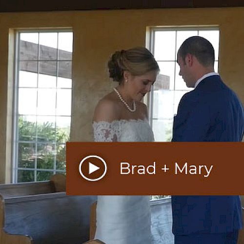 A couple in wedding attire stands close together in a chapel. Overlaid text: Brad + Mary, with a play button icon.