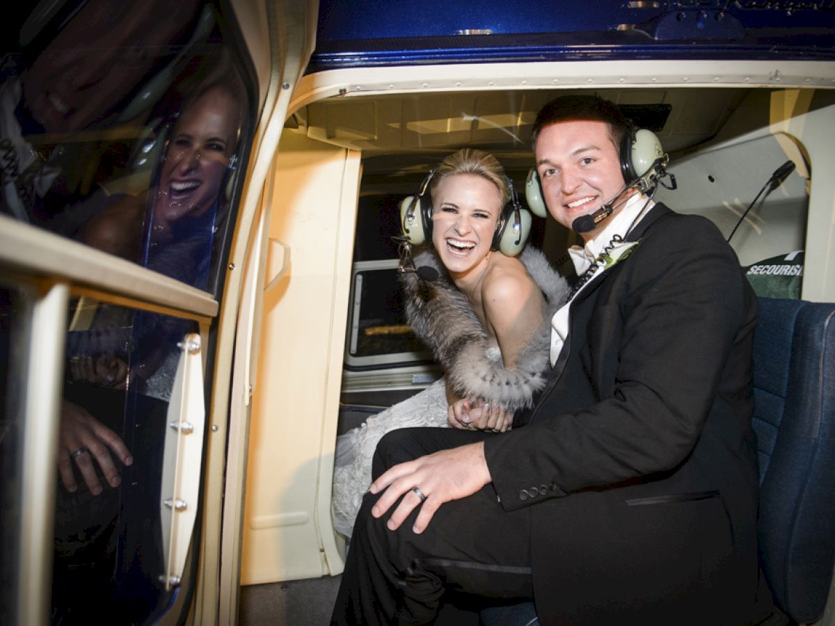 A couple in formal attire, both wearing headsets, smiles inside a helicopter with a reflection visible on the window.